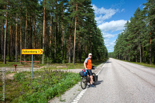 Cyclist standing by a road sign in Sweden, surrounded by tall pine trees and clear summer sky during a long‑distance bike trip
