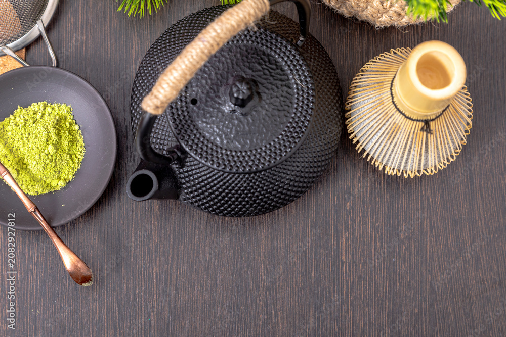 Fototapeta premium Matcha tea powder with bamboo whisk and black iron teapot on dark wooden table. Japanese tea ceremony preparation with organic green tea leaves. Healthy natural lifestyle tradition