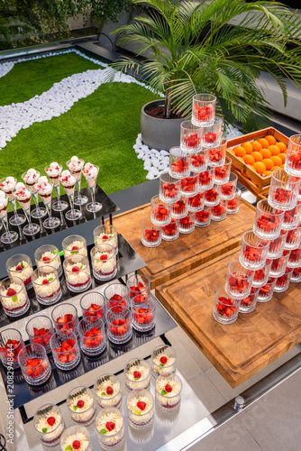 High-angle view of an elegant outdoor catering dessert station featuring a pyramid of strawberry cups, creamy trifles, and a wooden crate of fresh oranges on a manicured garden terrace.
