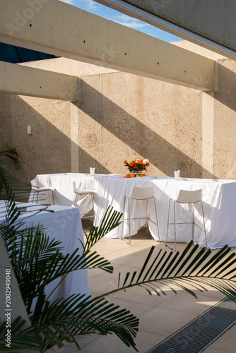 Minimalist outdoor event setup with white covered high tables and chairs under concrete beams, featuring a vibrant floral centerpiece with oranges on a modern terrace with architectural shadows.