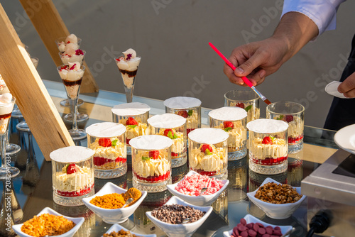 Chef's hand carefully decorating individual creamy dessert trifles with toppings in a glass station, featuring various bowls of garnishes like chocolate, nuts, and sprinkles at a catering event.