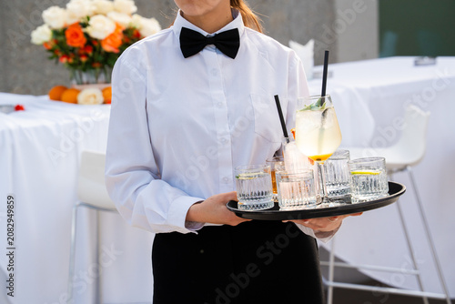 A professional waiter in a white shirt and black bow tie serving assorted cocktails and water in elegant glasses on a black tray at a formal outdoor catering event.