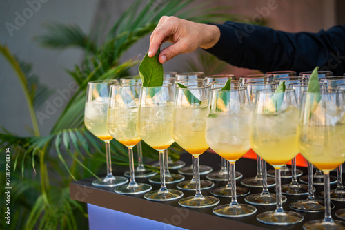 Hand of a bartender carefully garnishing a row of refreshing yellow cocktails in wine glasses with fresh green sage leaves at an upscale outdoor catering event.
