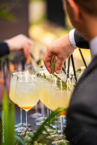 Hand of a bartender carefully garnishing a row of refreshing yellow cocktails in wine glasses with fresh green sage leaves at an upscale outdoor catering event.