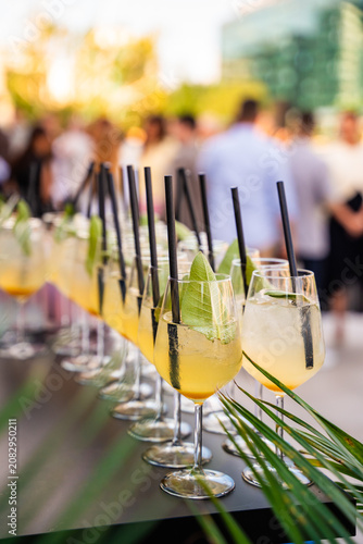 Close-up of a row of refreshing cocktails with black straws and sage leaf garnishes on a service table at a crowded outdoor summer event with a blurred background.