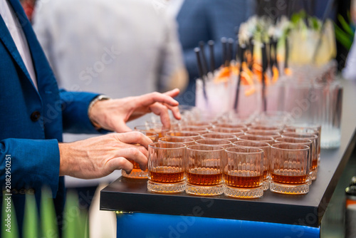 A person in a blue suit adjusting crystal glasses filled with amber-colored whiskey or spirit on a service table at an upscale catering event, with blurred party guests in the background.