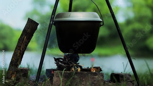 Metal pot suspended over campfire by tripod cookout outdoors. Cooking activity with smoke, fire, and glowing embers against blurred green background of river and trees.