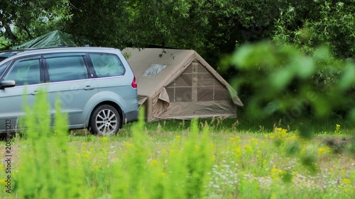 Light blue minivan vehicle parked near large tan canvas camping tent setup in lush green field, abundant trees and wildflowers surround outdoor summer recreation area, copy space