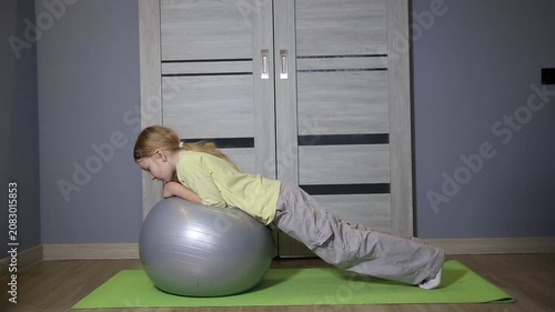 Young girl balancing on large exercise ball performing plank exercise in home room, engaging core muscles for fitness and strength training, developing stability and physical condition.
