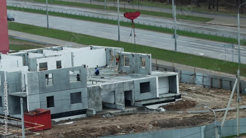Construction of multistory building using concrete panels by construction workers under large red crane hook overhead, urban development project next to highway road infrastructure.