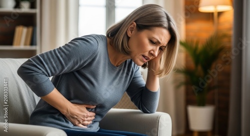 Woman suffering from severe stomach ache and abdominal pain while sitting on a couch at home.