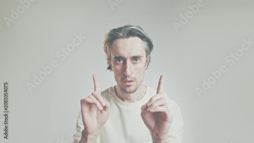 Serious young man in white sweater raising both index fingers, emphasis gesture, isolated on neutral background, communication and presentation concept