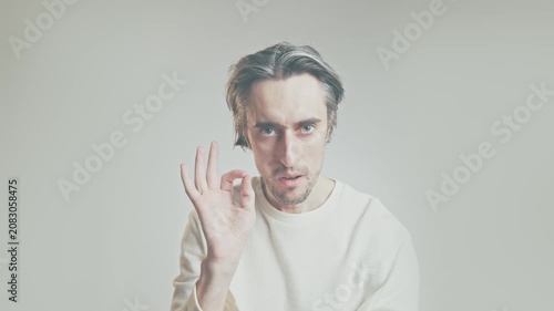 Serious adult man making OK hand gesture, looking at camera, light sweater, isolated on plain light background, approval and consent concept