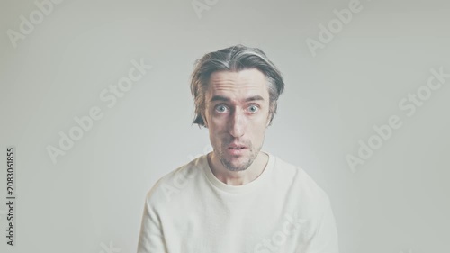 Surprised young man portrait with medium-length hair and light stubble, wearing long-sleeve shirt, in soft gray studio background, confused expression