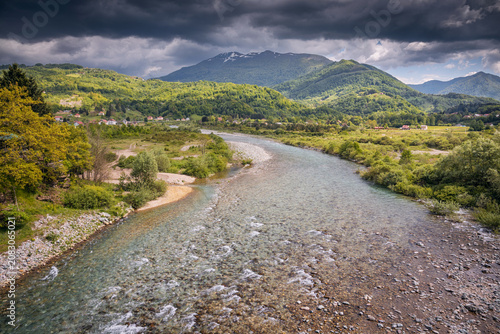 River with clear water running through a lush valley, surrounded by green hills and distant mountains under dark storm clouds