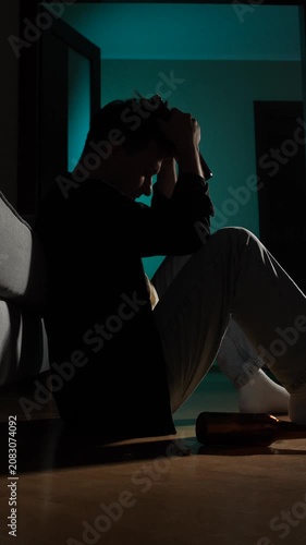 Vertical video silhouette of a man sitting on the floor in a dark room, holding a bottle to his head, depicting struggles with depression, addiction, and loneliness