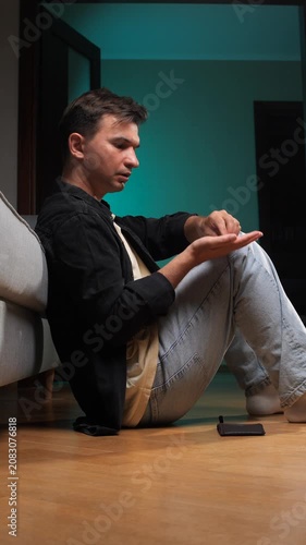 Vertical video young man sitting on the floor, holding an empty wallet over his open palm, contemplating financial problems and the lack of money