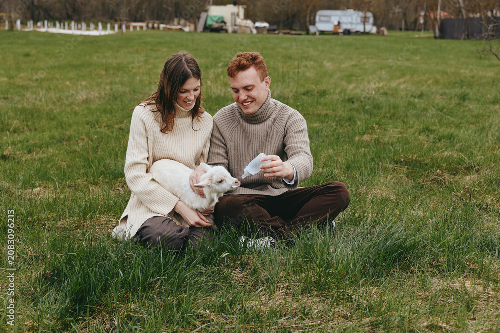 Fototapeta premium Couple sitting on green grass in a park, a white kid goat between them, sharing a gentle moment of care and play, cozy sweaters, casual lifestyle, and rustic charm.