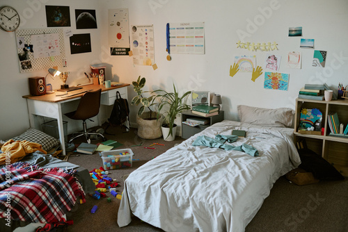 Wide angle view of messy child bedroom showing study desk, unmade bed, books, toys, drawings, signaling busy family routine and home organization. Ideal for parenting, education, storage marketing