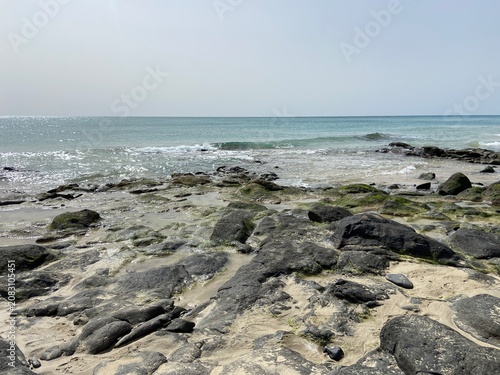 Rocky volcanic shoreline on the coast of Fuerteventura in the Canary Islands, Spain. Rugged black lava rocks meet the clear Atlantic Ocean beneath a bright blue sky, showcasing the island's dramatic 
