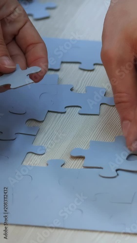A woman is assembling a gray puzzle on a wooden table. The puzzle pieces are scattered, and the woman carefully places a gray piece into the emerging gray shape.
