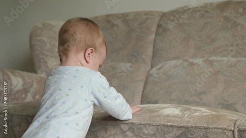 Baby practices standing on soft furniture. Curious infant knocks on the sofa. Young child experiments with standing by sofa in cozy environment with sunlight