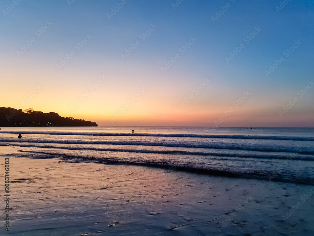 Fototapeta premium Tropical beach at twilight with orange and blue sunset sky, silhouetted headland and fishing boat on calm ocean