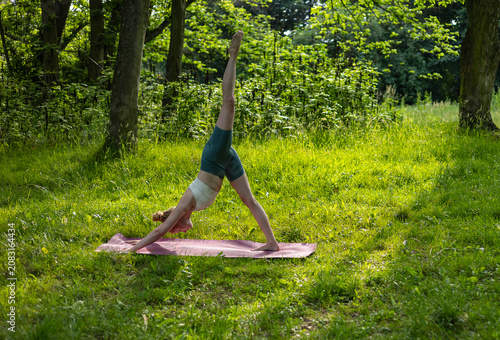 Young fit woman doing a three legged downward facing dog yoga pose on a mat in a sunny green forest clearing. Active healthy lifestyle, outdoor training and body flexibility exercise