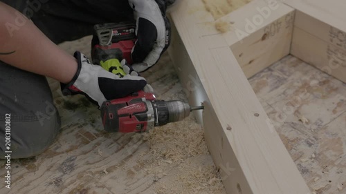 Close-up of worker fastening a wooden frame with a cordless drill on a construction floor with wood shavings