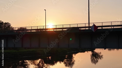 Cars driving across a bridge at sunset, with reflections visible in the water below and trees lining the road, creating a serene evening atmosphere