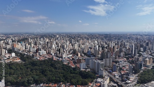 Foto Visão aérea da cidade de Campinas, SP, com prédios altos, bairros densos, áreas verdes e horizonte urbano amplo sob céu azul, mostrando a expansão da metrópole no interior paulista