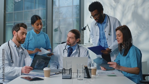 Medical team cooperating working together in clinic. Caucasian doctor boss leader confident talk leading medicine conference with multiethnic five diverse people doctors nurses at meeting in hospital