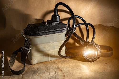 Vintage miner's headlamp with heavy battery pack glowing on retro background. Retro industrial mining lantern with cable and strap. Soviet underground equipment. Speleology