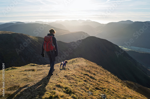 A female hiker and their dog walking down from the summit of Grasmoor with Whiteless Pike and Robinson in the distance on a bright winters morning in the Lake District, UK