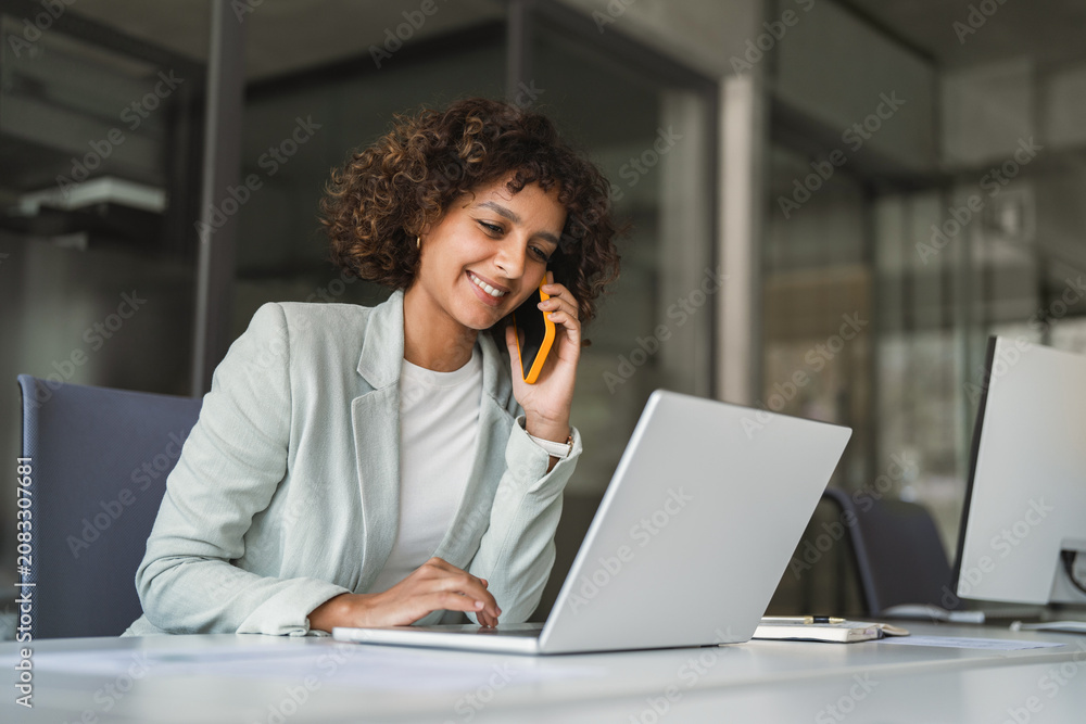 Fototapeta premium Young Indian woman manager ceo using cell phone mobile, laptop for business call. Smiling Latin female businesswoman holding smartphone in office, talking with client, doing professional consultation