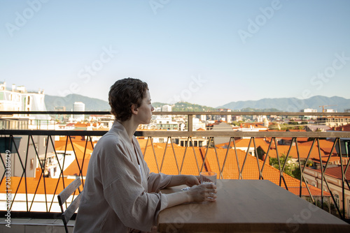 woman recovering from cancer looks across the city from a balcony table with water and medicine, conveying survivorship, quiet reflection and ongoing healthcare