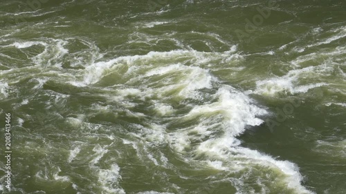 Raging green water of a powerful river creating waves and white foam. Turbulent stream representing nature's force and untamed environment