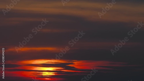 Airplane flying high in evening sky above dramatic sunset with glowing red sun and dark clouds, static telephoto shot