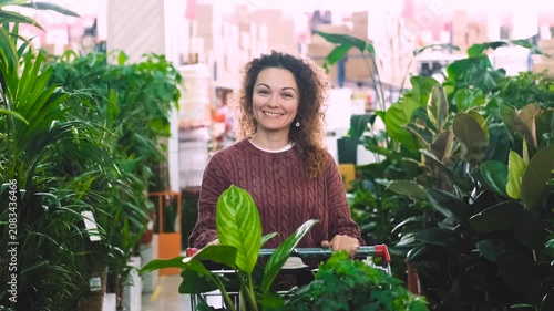 A happy woman with curly hair buys potted flowers at a garden center. The woman carries the flowers in a shopping cart.