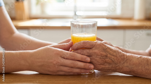 Caregiver providing support to elderly person, young hands holding glass of orange juice together with senior hands reflecting care