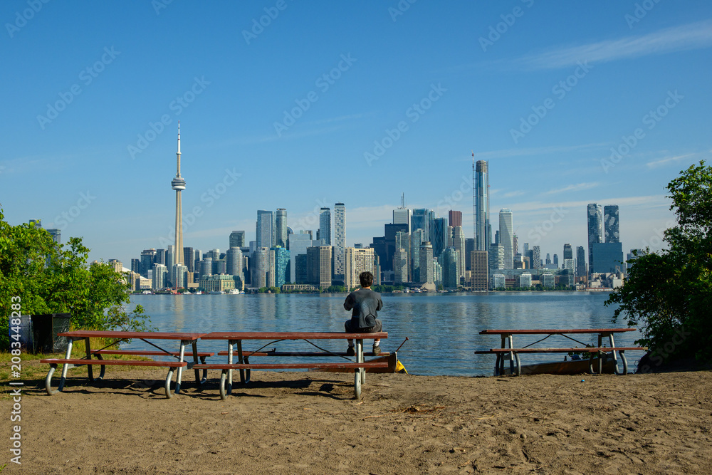 Obraz premium Person sitting on a picnic table photographing the Toronto skyline from a sandy island shore.