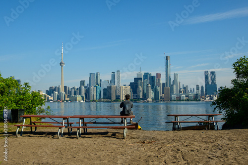Person sitting on a picnic table photographing the Toronto skyline from a sandy island shore.