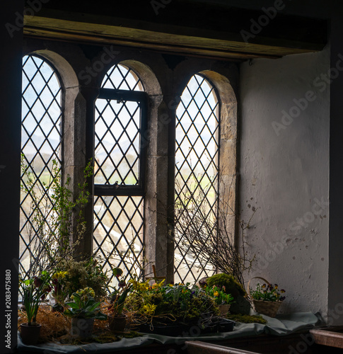 A traditional triple-arched window with stone mullions and leaded glazing, adorned with floral arrangements and potted plants on the sill inside the church