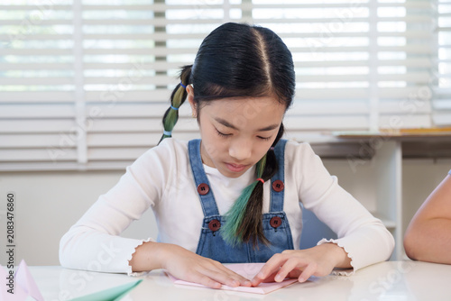 Photos Asian preteen girl carefully folding pink origami paper during classroom craft a