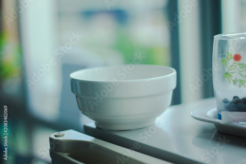 A minimalist white ceramic bowl placed on a digital kitchen scale beside a clear glass, illuminated by soft natural window light. A clean, modern kitchen scene with a calm atmosphere, perfect for food