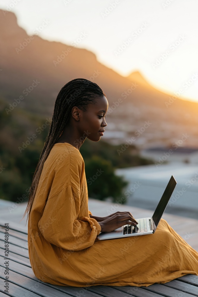 Naklejka premium Young woman on a deck using laptop at sunset in a yellow dress for remote work background and website banner design