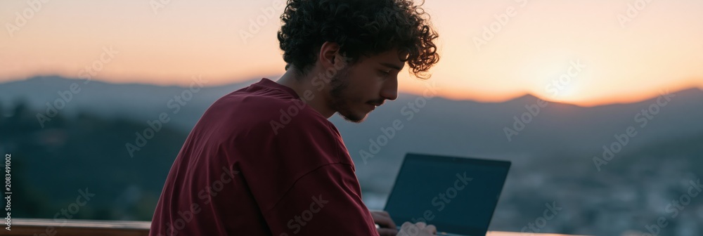 Fototapeta premium Young man with laptop at sunset on a terrace overlooking hills for remote work background and banner design