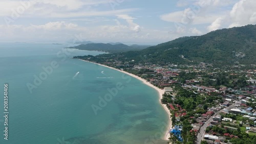 A sandy beach lined with tropical greenery and turquoise waters under a bright sky. Ko Samui, Thailand.