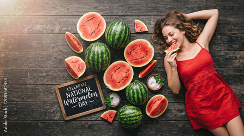 Beautiful Woman in Red Dress Lying on Vintage Wooden Background and Eating Fresh Watermelon, National Watermelon Day Concept