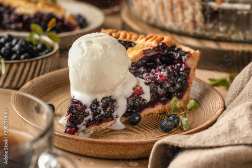 Slice of blueberry pie with a scoop of ice cream on ceramic plate on brown textured table background, traditional homemade dessert